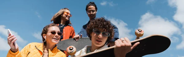 Vue à faible angle des patineurs interraciaux près d'amis joyeux contre le ciel nuageux bleu, bannière — Photo de stock