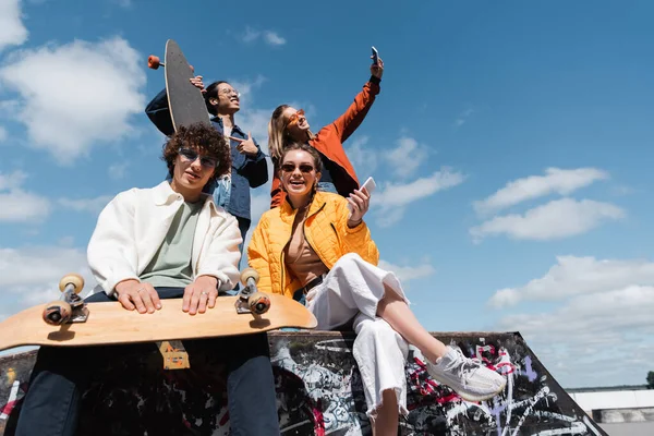 Bajo ángulo vista de amigos mirando cámara cerca de mujer tomando selfie con asiático skater - foto de stock