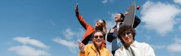 Happy woman taking selfie with asian skater and friends against blue sky, banner — Stock Photo