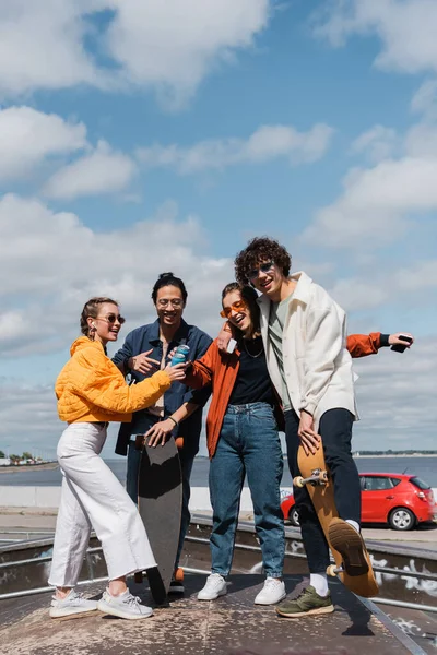 Amigos alegres tintineo latas de refrescos cerca de patinadores interracial al aire libre - foto de stock