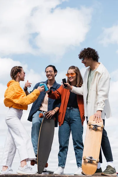 Happy young women clinking soda cans near trendy interracial skaters — Stock Photo