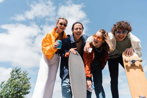 Vista de ángulo bajo de patinadores interracial cerca de amigos riendo con teléfonos inteligentes y latas de refrescos contra el cielo nublado - foto de stock