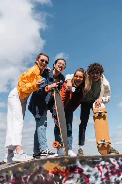 Volle Länge der asiatischen Skater zeigt Rock-Zeichen in der Nähe fröhlicher Freunde vor blauem bewölkten Himmel — Stockfoto