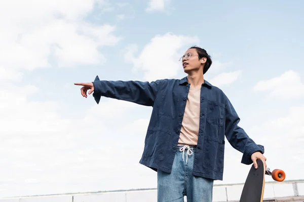 Young asian man with longboard looking away and pointing with finger outdoors — Stock Photo