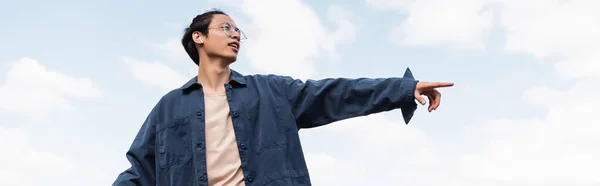 Baixo ângulo vista de asiático homem no óculos e fone de ouvido apontando com dedo ao ar livre, banner — Fotografia de Stock