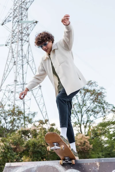 Visão de baixo ângulo do jovem homem encaracolado no skate jaqueta branca no parque de skate — Fotografia de Stock