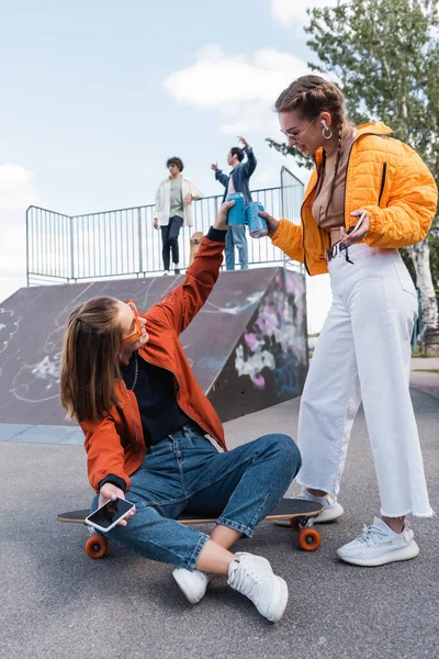 Mujeres elegantes latas de refrescos tintineo cerca de amigos en la rampa de skate sobre fondo borroso - foto de stock