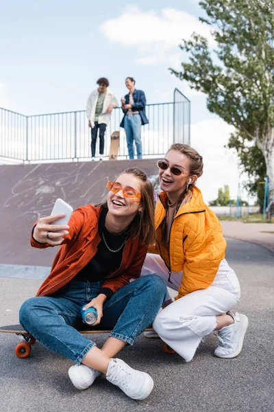 Donne gioiose scattare selfie in skate park vicino amici sulla rampa su sfondo sfocato — Foto stock