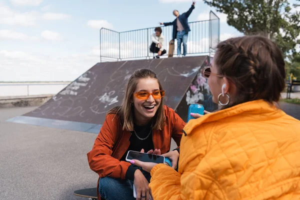 Mujer sonriente en gafas de sol con estilo sentado cerca de amigo y los hombres borrosos en la rampa de skate - foto de stock