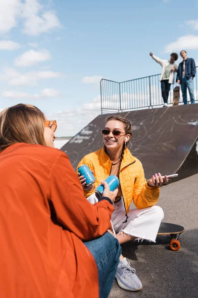 Улыбающаяся женщина в очках разговаривает с другом рядом с размытыми фигуристами на рампе в скейт-парке — стоковое фото