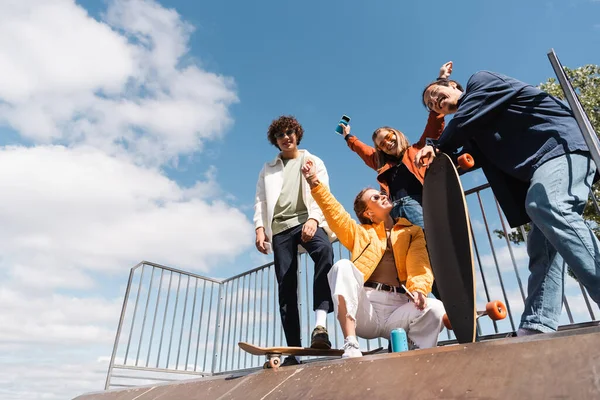 Vista de ángulo bajo de amigos multiétnicos alegres en la rampa de skate contra el cielo azul nublado - foto de stock