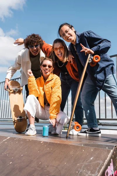 Cheerful women sticking out tongue near interracial skaters on ramp in skate park — Stock Photo
