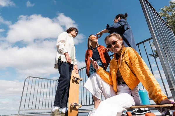 Mujer feliz con teléfono inteligente mirando a la cámara cerca de amigos contra el cielo nublado - foto de stock