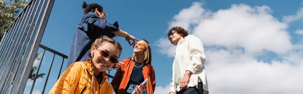 Vue à angle bas de la jeune femme souriant à la caméra près d'amis interraciaux contre ciel nuageux, bannière — Photo de stock