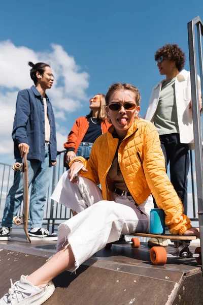 Жінка в сонцезахисних окулярах стирчить язиком, сидячи на скейтборді біля міжрасових друзів — стокове фото