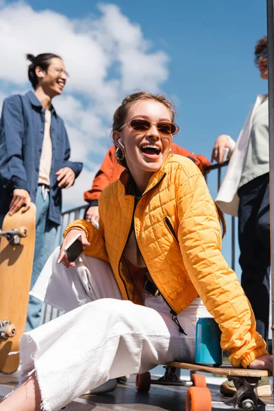 Mujer sorprendida en gafas de sol sentada cerca de patinadores multiétnicos borrosos contra el cielo azul - foto de stock