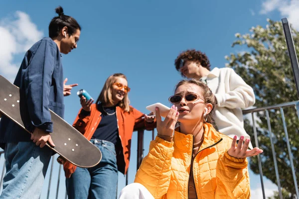 Mujer elegante que envía mensaje de voz en el teléfono inteligente cerca de patinadores interracial borrosa - foto de stock