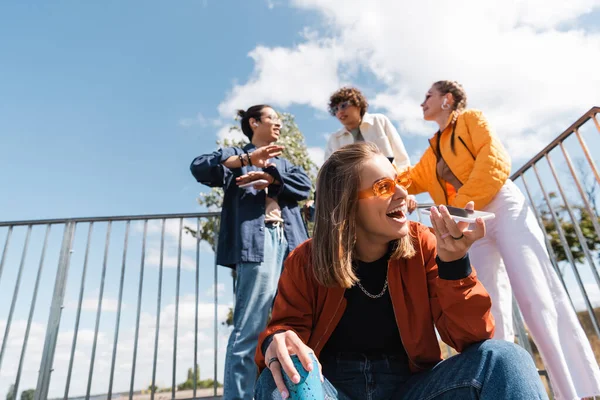 Femme excitée dans des lunettes de soleil envoyer un message vocal près d'amis multiethniques sur fond flou — Photo de stock