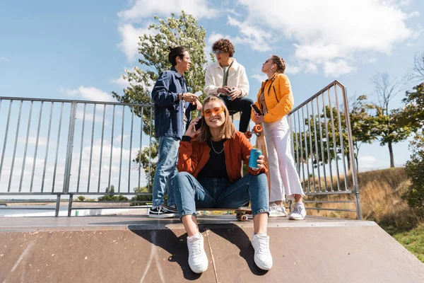 Mulher sorridente com lata de refrigerante sentado na rampa de skate, enquanto amigos multiculturais falando em segundo plano — Fotografia de Stock