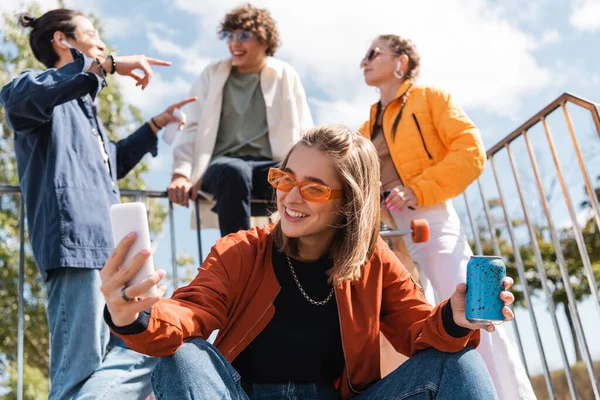 Mulher feliz com refrigerante pode tomar selfie perto de amigos multiétnicos no fundo borrado — Fotografia de Stock