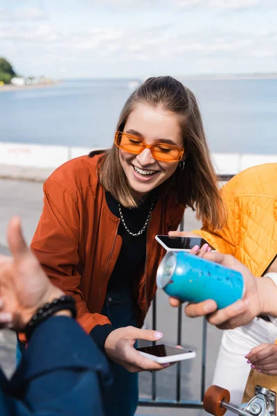 Улыбающаяся женщина в модных солнцезащитных очках держит смартфон с чистым экраном рядом с друзьями — стоковое фото