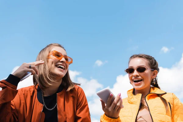 Mujer en gafas de sol con estilo celebración de teléfono inteligente cerca amigo feliz contra el cielo azul - foto de stock