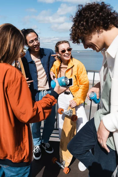 Patinadores interracial felices y elegantes con latas de refrescos hablando al aire libre - foto de stock