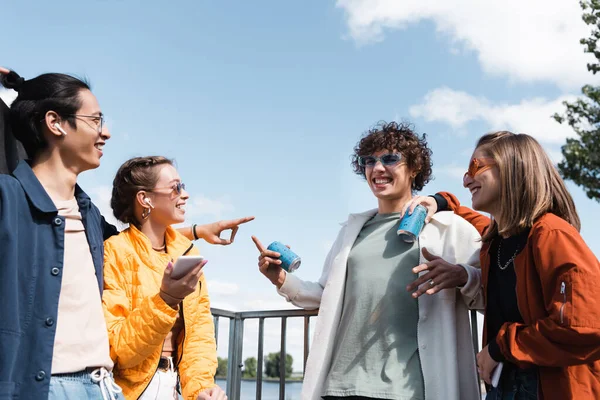 Amigos multiculturales alegres con latas de refrescos apuntando el uno al otro al aire libre — Stock Photo