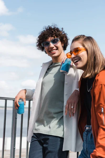 Jovem casal em roupas elegantes e óculos de sol de pé com latas de refrigerante ao ar livre — Fotografia de Stock