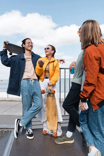 Happy asian man holding skateboard near friends with smartphones and soda can — Stock Photo