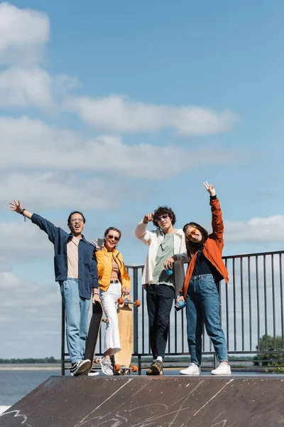 Heureux patineurs multiculturels agitant les mains et montrant des signes de victoire sur la rampe de patinage sous le ciel nuageux bleu — Photo de stock