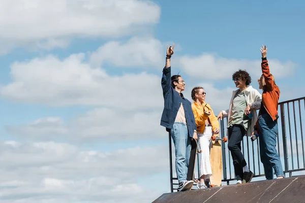 Alegre interracial amigos gesto en skate rampa bajo azul y nublado cielo - foto de stock