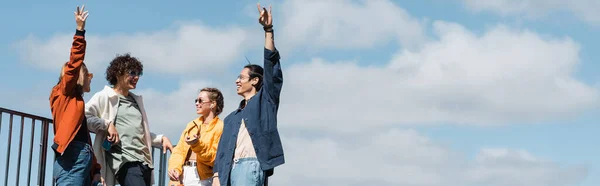 Щасливий азіатський чоловік, що показує скельний знак біля друзів проти блакитного хмарного неба, банер — стокове фото