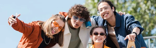 Mujer alegre con teléfono inteligente sobresaliendo lengua cerca de amigos multiétnicos, pancarta - foto de stock