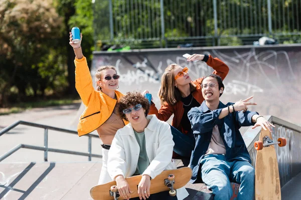 Jovem feliz com lata de refrigerante na mão levantada perto de amigos multiétnicos no parque de skate — Fotografia de Stock