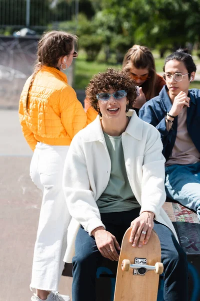 Hombre rizado con monopatín mirando a la cámara cerca de amigos multiculturales - foto de stock