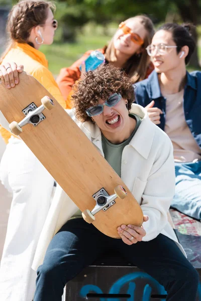 Curly man with skateboard grimacing at camera near blurred multiethnic friends — Stock Photo