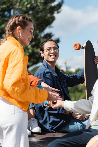 Feliz asiático hombre en gafas celebración longboard cerca amigos al aire libre — Stock Photo