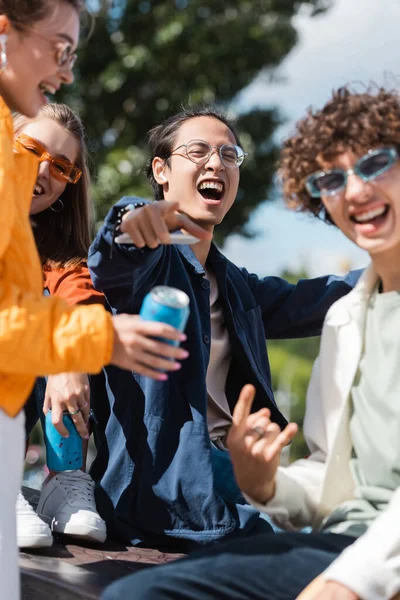 Excited asian man in eyeglasses pointing with finger near blurred friends outdoors — Stock Photo