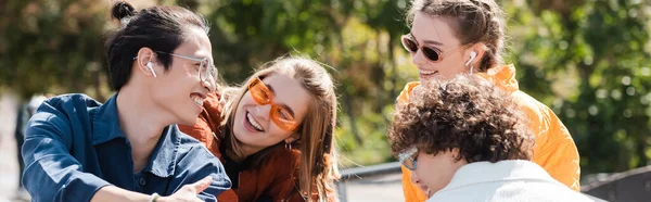 Asiático hombre en inalámbrico auricular sonriendo cerca feliz amigos al aire libre, bandera - foto de stock