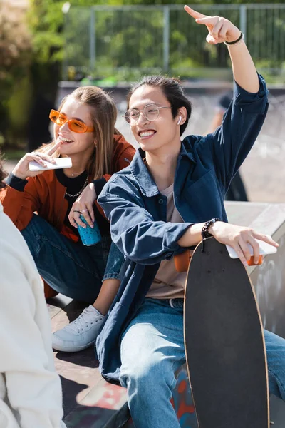 Sorridente asiatico uomo con longboard mostrando vittoria segno vicino amici all'aperto — Foto stock