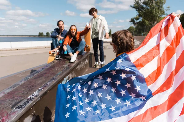 Femme tenant le drapeau des Etats-Unis près des amis multiethniques flous dans le skate park — Photo de stock