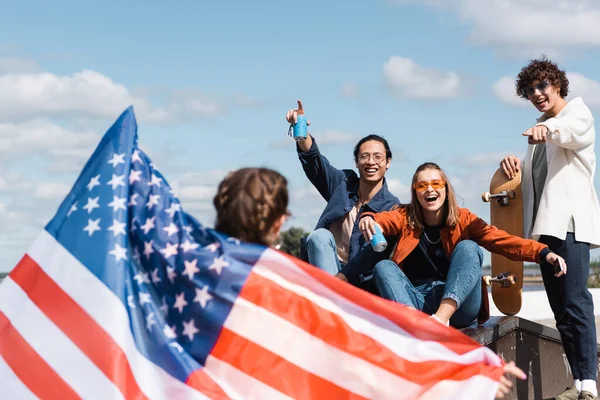 Fröhliche multiethnische Freunde zeigen auf verschwommene Frau mit US-Flagge im Freien — Stockfoto