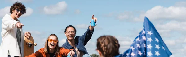 Smiling skater pointing at woman with usa flag near interracial friends with soda cans, banner — Stock Photo