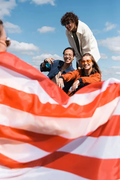 Smiling interracial friends looking at woman with usa flag on blurred foreground — Stock Photo