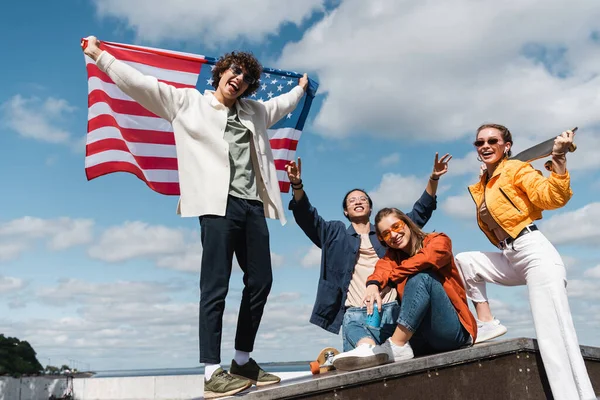 Homem encaracolado segurando bandeira EUA perto de amigos multiétnicos alegres — Fotografia de Stock