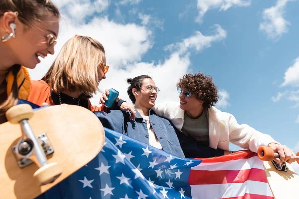 Patinadores multiétnicos felizes com bandeira dos EUA abraçando o céu azul e nublado — Fotografia de Stock
