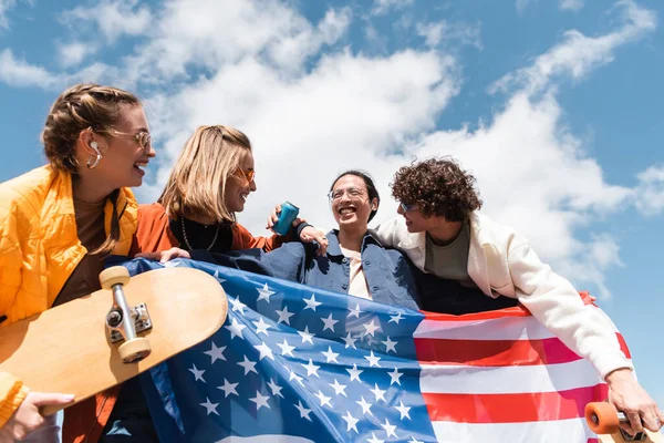 Joyful multicultural skaters holding usa flag under blue cloudy sky — Stock Photo