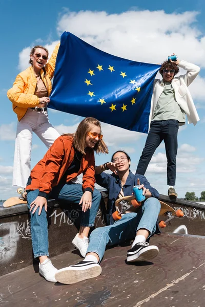Alegres amigos multiétnicos con bandera de la unión europea en skate park - foto de stock