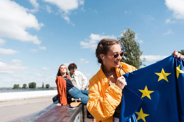 Mujer en gafas de sol de moda sonriendo mientras sostiene la bandera de la UE cerca de amigos borrosos - foto de stock